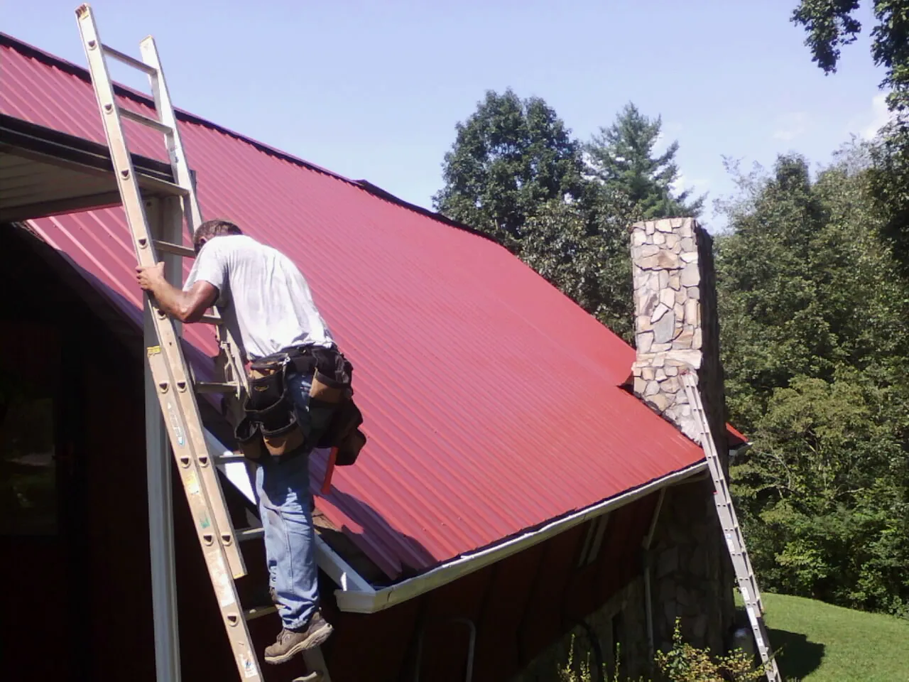 Roofer on a ladder beside a red metal roof and stone chimney — Soffit & Fascia Repair by Roofing Brothers in Medical Lake